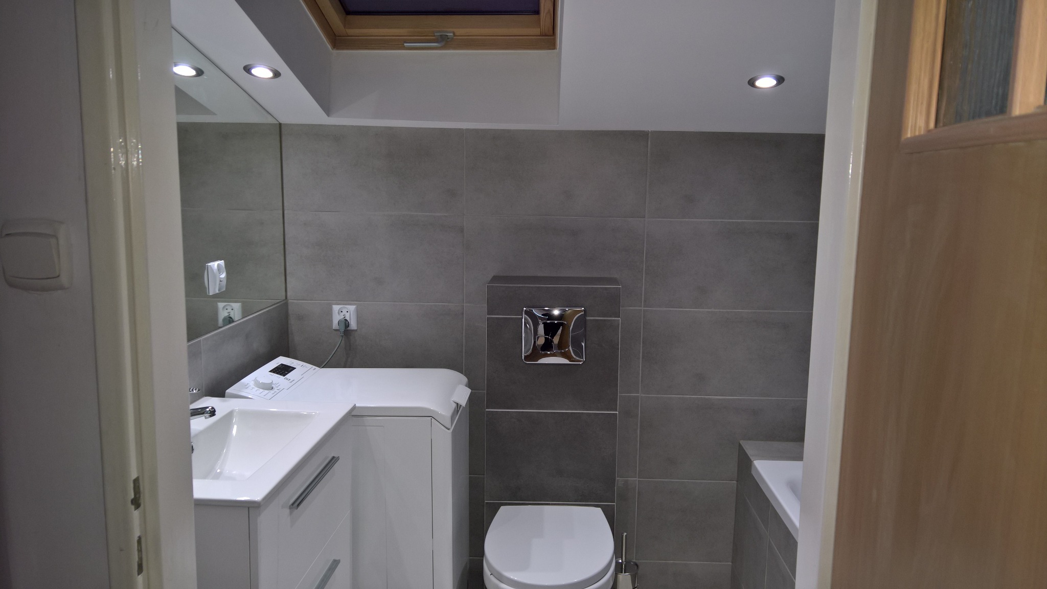 Nowoczesna łazienka w szarościach z pralką zabudowaną pod umywalką, wisząca toaleta z chromowanym przyciskiem, okno dachowe w suficie.