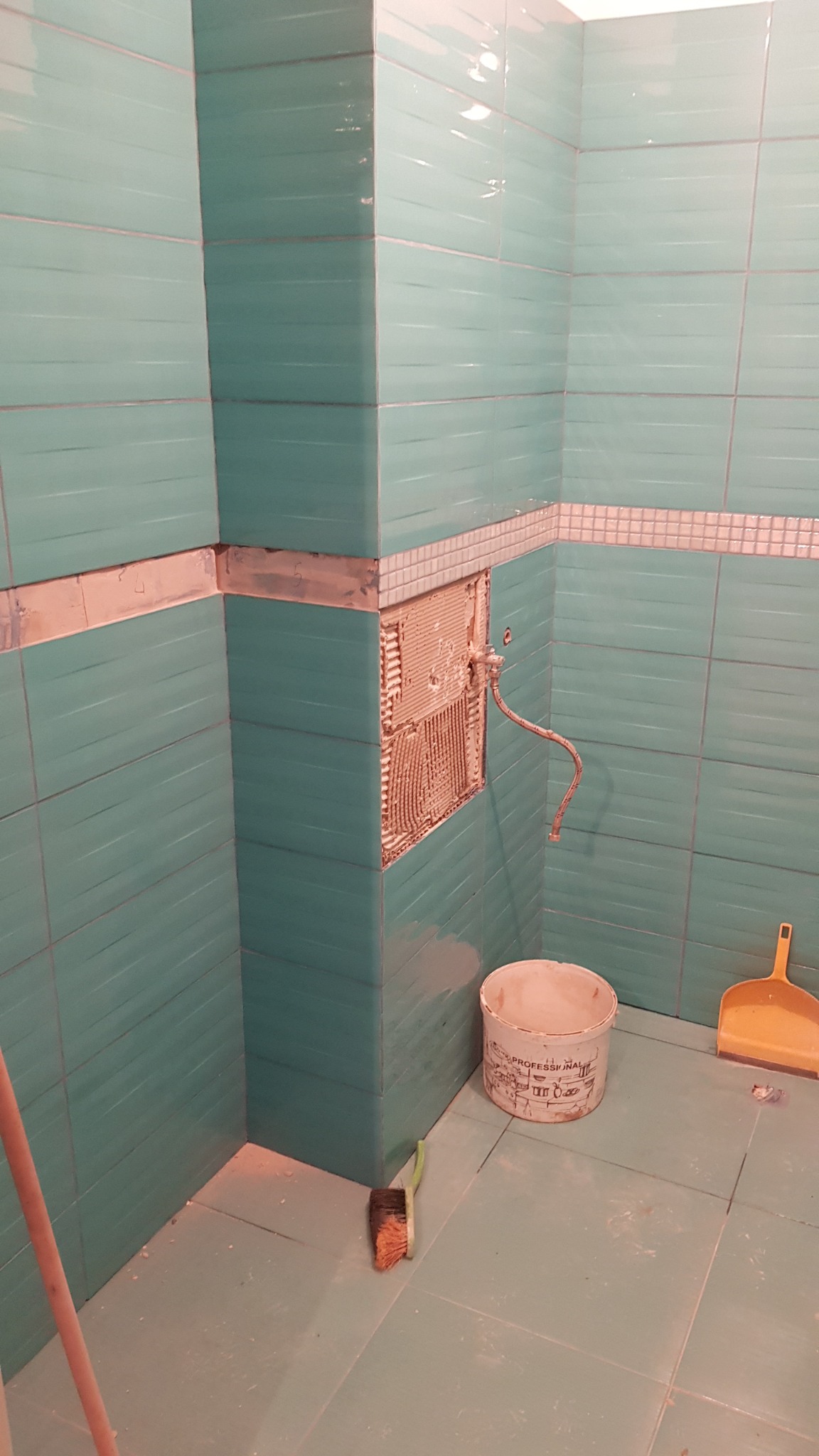 Niedokończona ściana w łazience z turkusowymi płytkami i odsłoniętą instalacją hydrauliczną, widoczne narzędzia i wiadro.