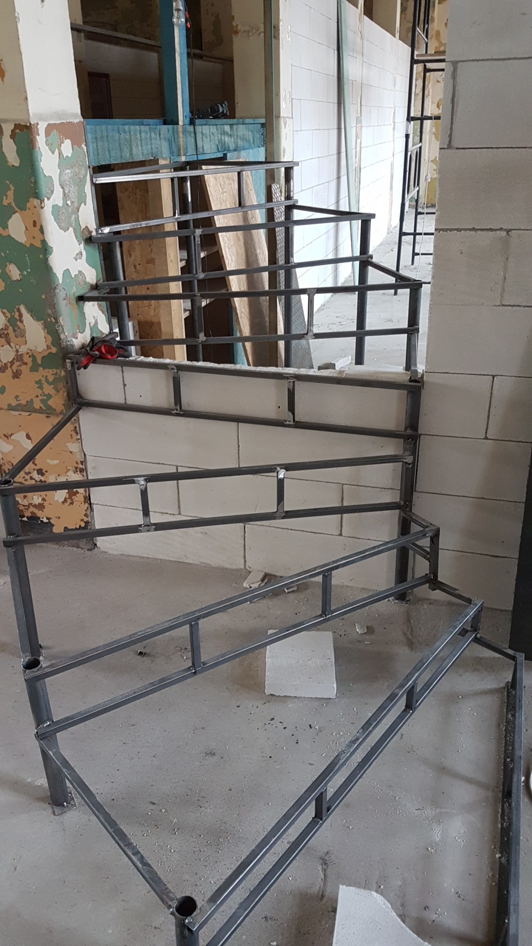 Metalowa konstrukcja schodów w surowym stanie, przymocowana do ściany z bloków betonowych, widoczne ślady farby na ścianie w tle, czerwone rękawice robocze na górnym poziomie konstrukcji.