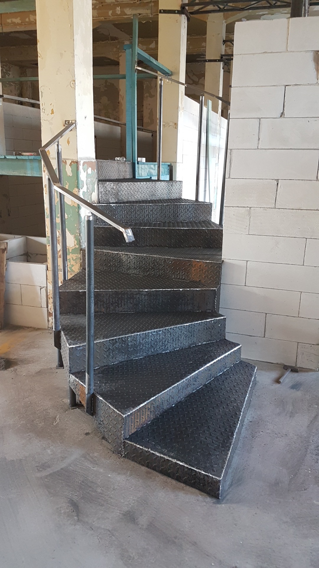 Metalowe schody z blachy ryflowanej z poręczą, zamontowane w surowym wnętrzu budynku w trakcie remontu, widoczne ślady prac budowlanych i fragmenty ścian z betonu komórkowego.