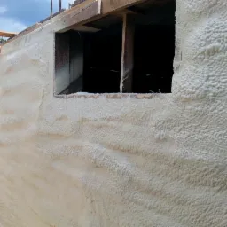 Ściana fundamentowa budynku w trakcie izolacji natryskową pianą poliuretanową, widoczne drewniane elementy konstrukcyjne okna i zbrojenie stropu.