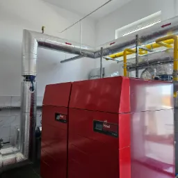 Czerwony piec gazowy Hoval UltraGas z izolowanymi rurami, widoczne żółte rury gazowe i srebrne rury instalacji grzewczej, białe ściany i szara podłoga z płytek.