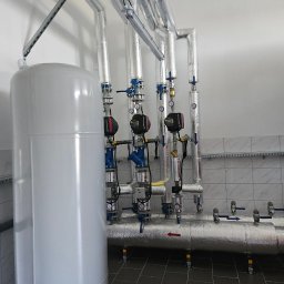 Z.U.H.Instalacje Sanitarne Grzegorz Stachowicz - Instalacja hydrauliczna w pomieszczeniu technicznym: biały zbiornik buforowy, izolowane rury z zaworami i manometrami, pompy obiegowe Grundfos, szara podłoga z płytek, ściany wyłożone kafelkami.