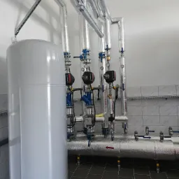 Instalacja hydrauliczna w pomieszczeniu technicznym: biały zbiornik buforowy, izolowane rury z zaworami i manometrami, pompy obiegowe Grundfos, szara podłoga z płytek, ściany wyłożone kafelkami.