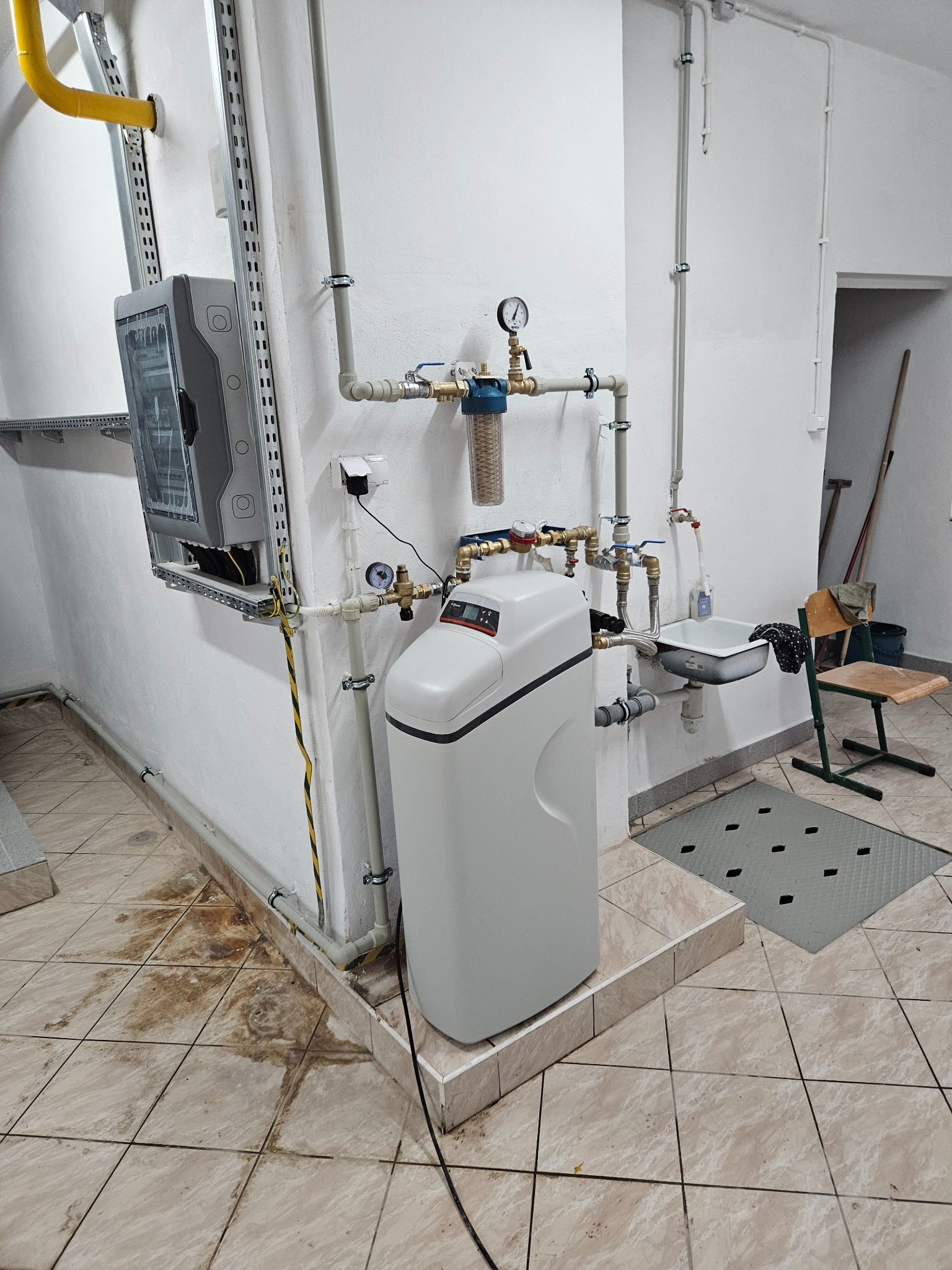 Biały zmiękczacz wody z manometrem zamontowany na ścianie w pomieszczeniu gospodarczym, widoczne rury, instalacja elektryczna, umywalka i kratka ściekowa na podłodze.