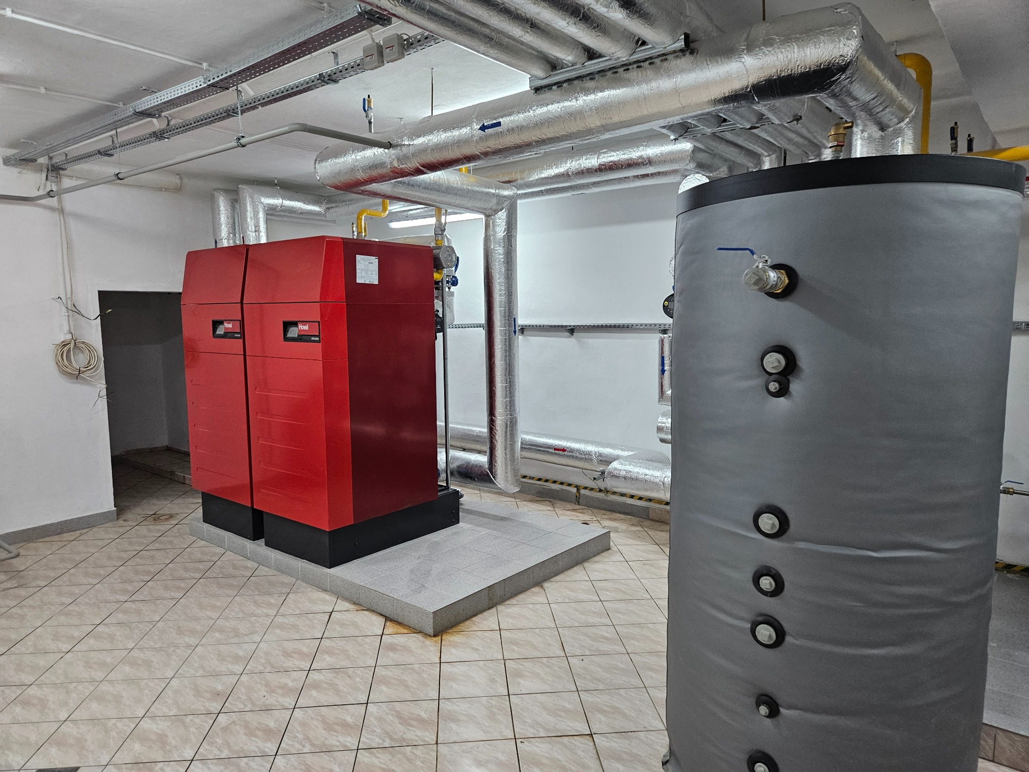 Czerwony piec grzewczy z szarym zbiornikiem akumulacyjnym w kotłowni, z widocznymi rurami instalacji grzewczej pod sufitem.