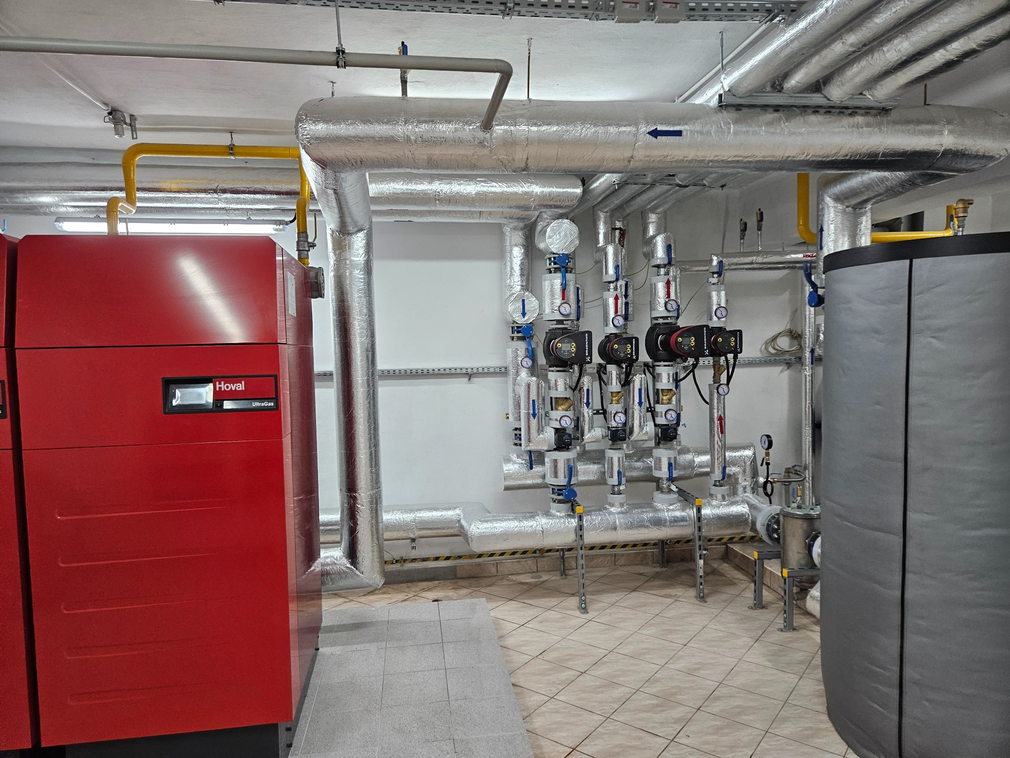 Kotłownia z czerwonym kotłem Hoval UltraGas, izolowanymi rurami, zaworami i manometrami. Rury oznaczono niebieską strzałką wskazującą kierunek przepływu. Podłoga wyłożona płytkami.