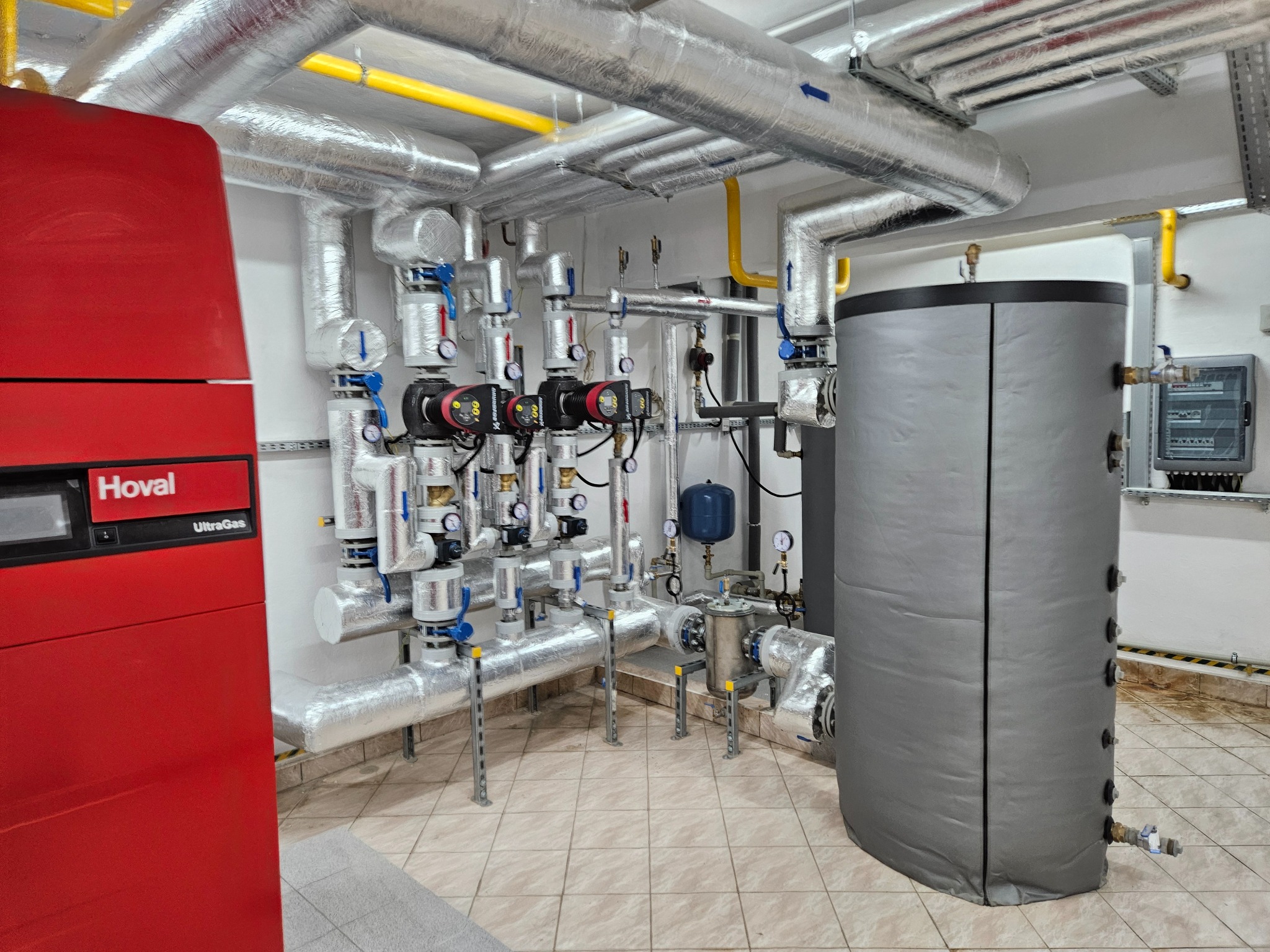 Kompleksowa kotłownia z czerwonym piecem Hoval UltraGas, zbiornikiem akumulacyjnym w szarej izolacji i licznymi rurami oraz pompami obiegowymi, zabezpieczonymi srebrną izolacją.