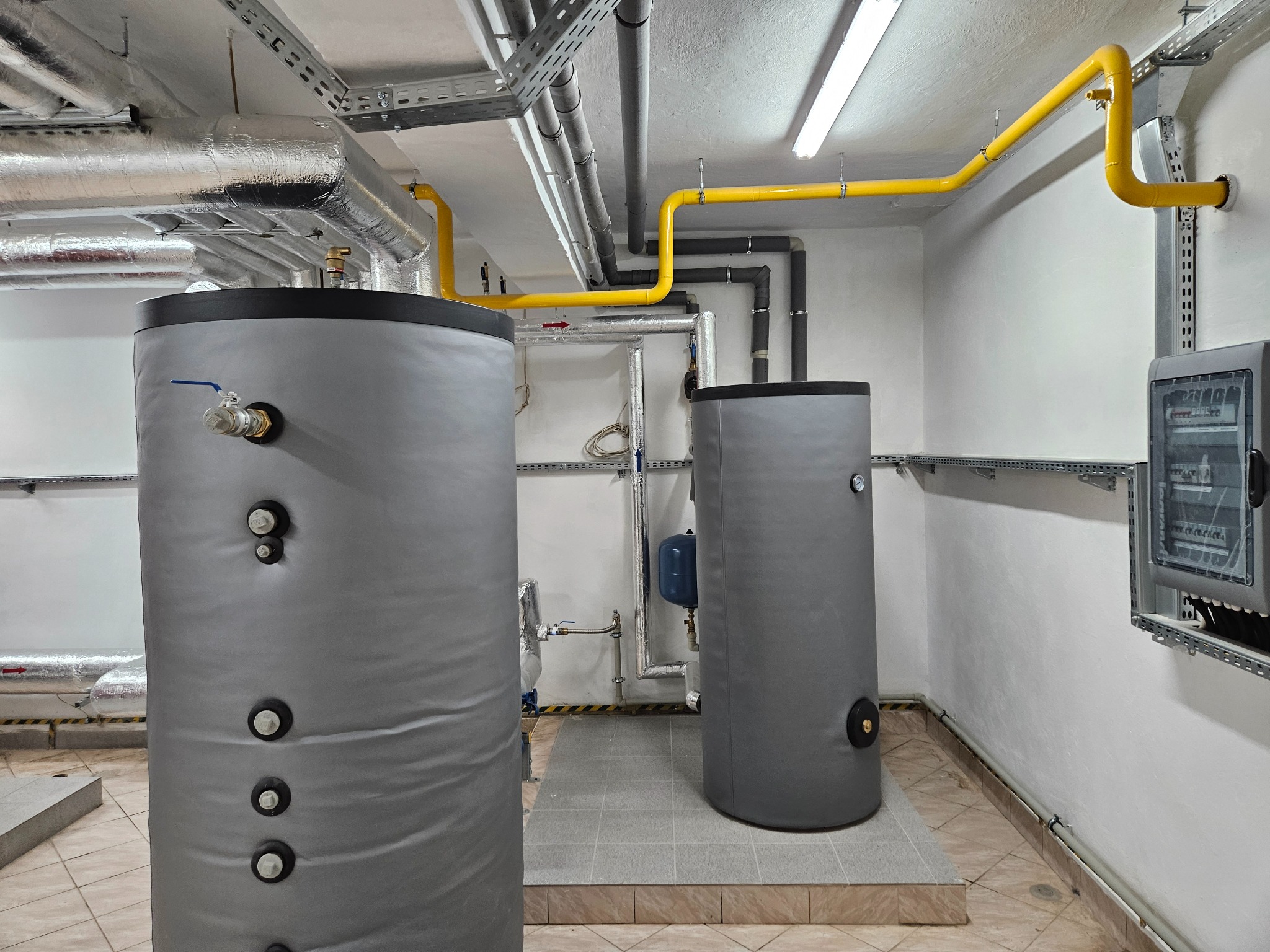 Dwa szare, cylindryczne zbiorniki buforowe w kotłowni, z widocznymi rurami w izolacji, żółtymi rurami gazowymi pod sufitem, oraz skrzynką elektryczną na ścianie.