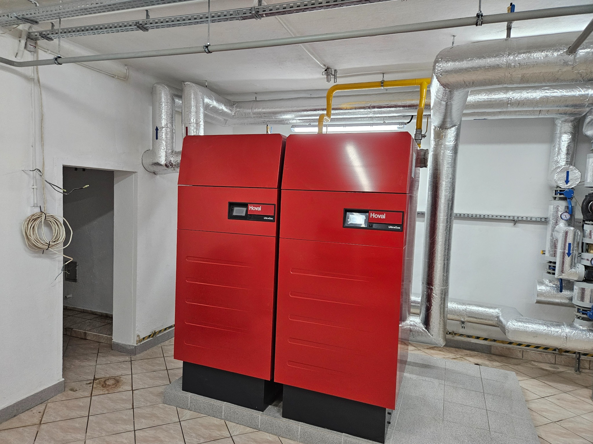 Dwa czerwone kotły grzewcze Hoval UltraGas w kotłowni, widoczne rury izolowane termicznie, instalacje pod sufitem i płytki podłogowe.