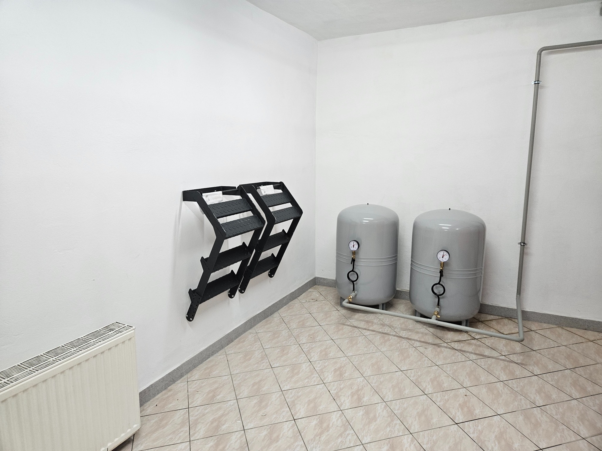 Dwa szare zbiorniki ciśnieniowe z manometrami i rurami w pomieszczeniu z białymi ścianami i beżową podłogą w kratkę, obok grzejnika i czarnych uchwytów naściennych.