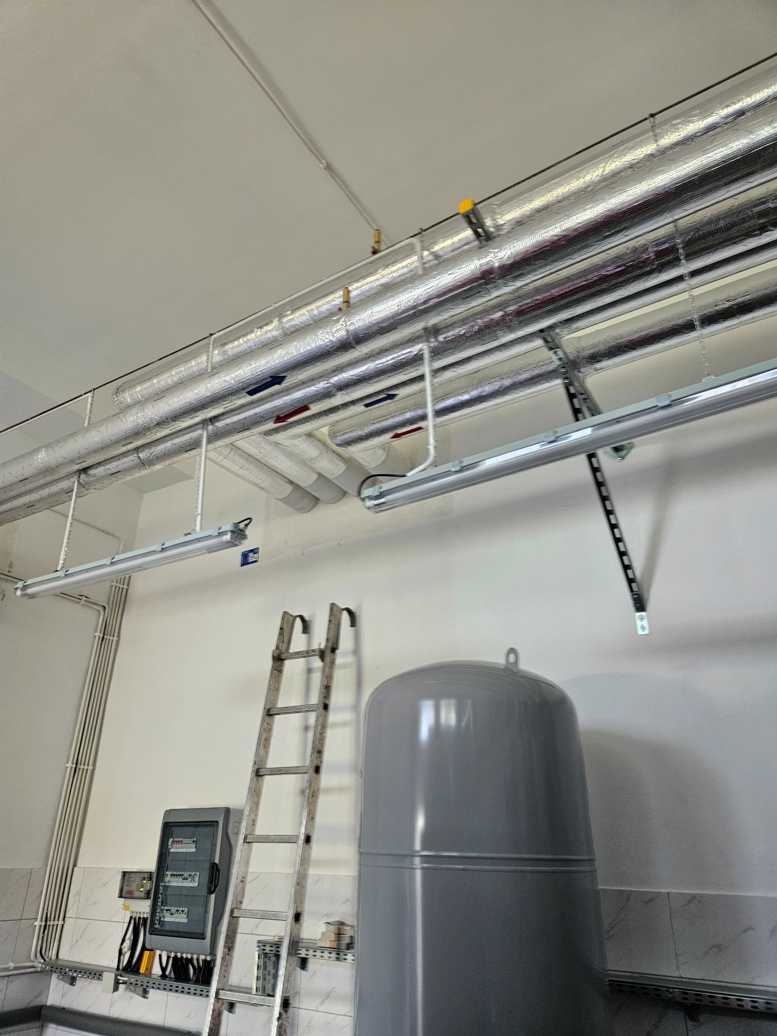 Szare naczynie przeponowe obok drabiny aluminiowej i skrzynki elektrycznej na tle białej ściany z rurami grzewczymi pod sufitem.