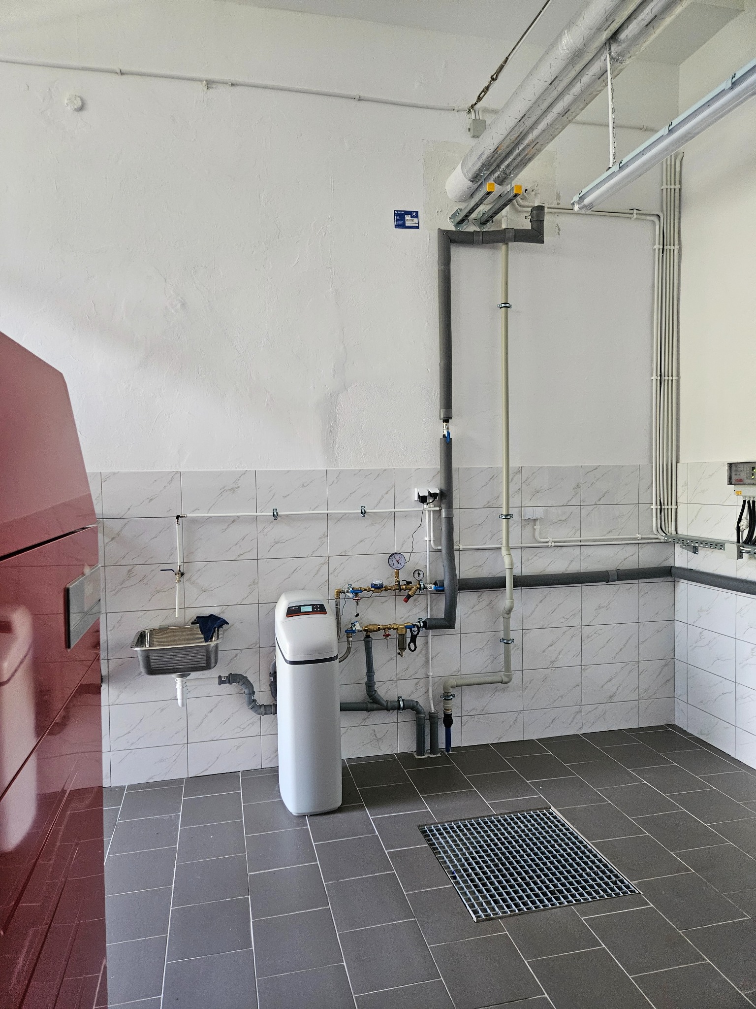 Wnętrze kotłowni z widocznym piecem, zmiękczaczem wody, instalacją hydrauliczną z rurami i zaworami, umywalką oraz kratką ściekową w podłodze.