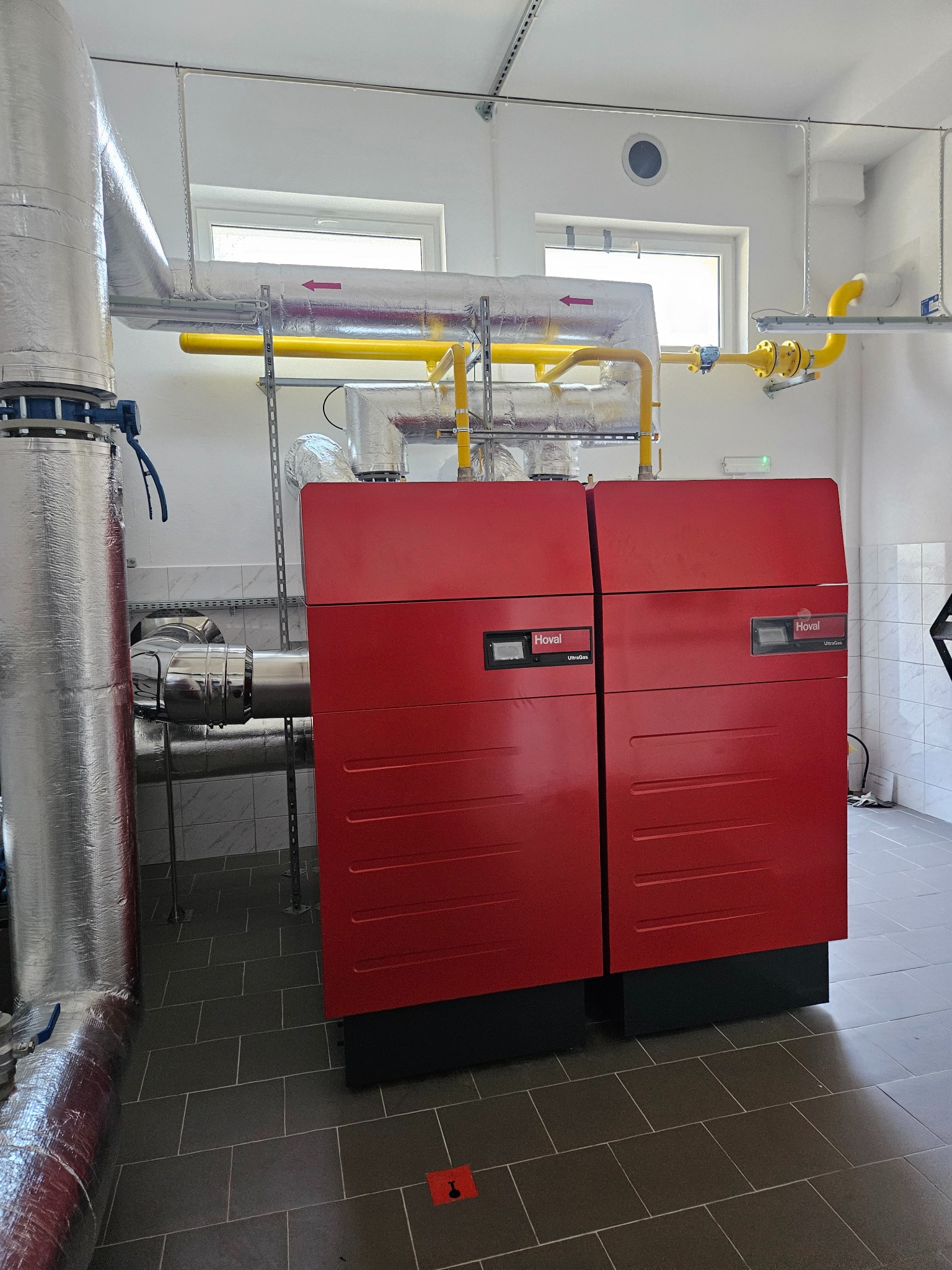 Czerwony piec gazowy marki Hoval UltraGas z widocznymi rurami gazowymi w kolorze żółtym i rurami izolowanymi termicznie, zamontowany w pomieszczeniu z szarą podłogą z płytek.