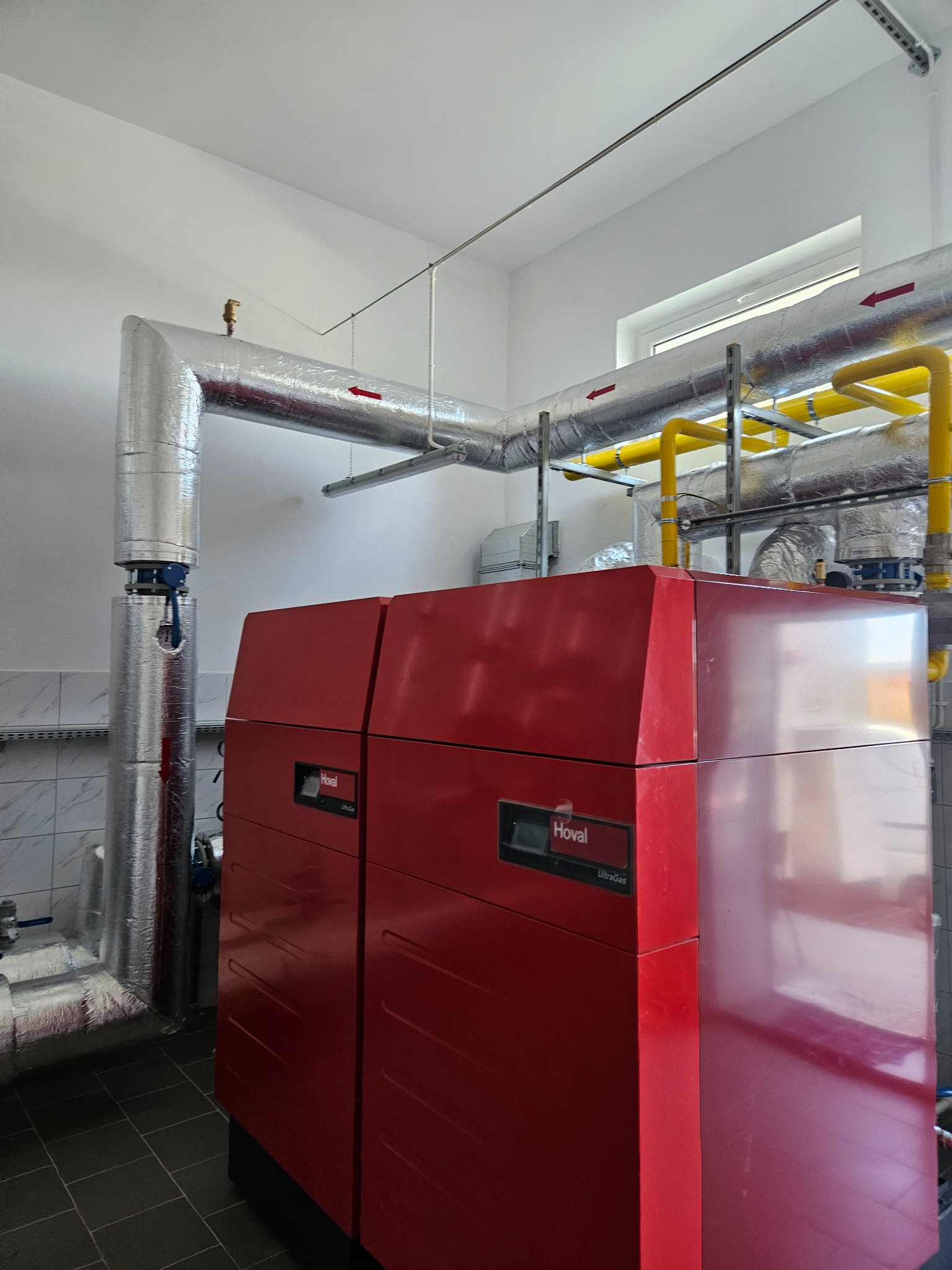 Czerwony piec gazowy Hoval UltraGas z izolowanymi rurami, widoczne żółte rury gazowe i srebrne rury instalacji grzewczej, białe ściany i szara podłoga z płytek.