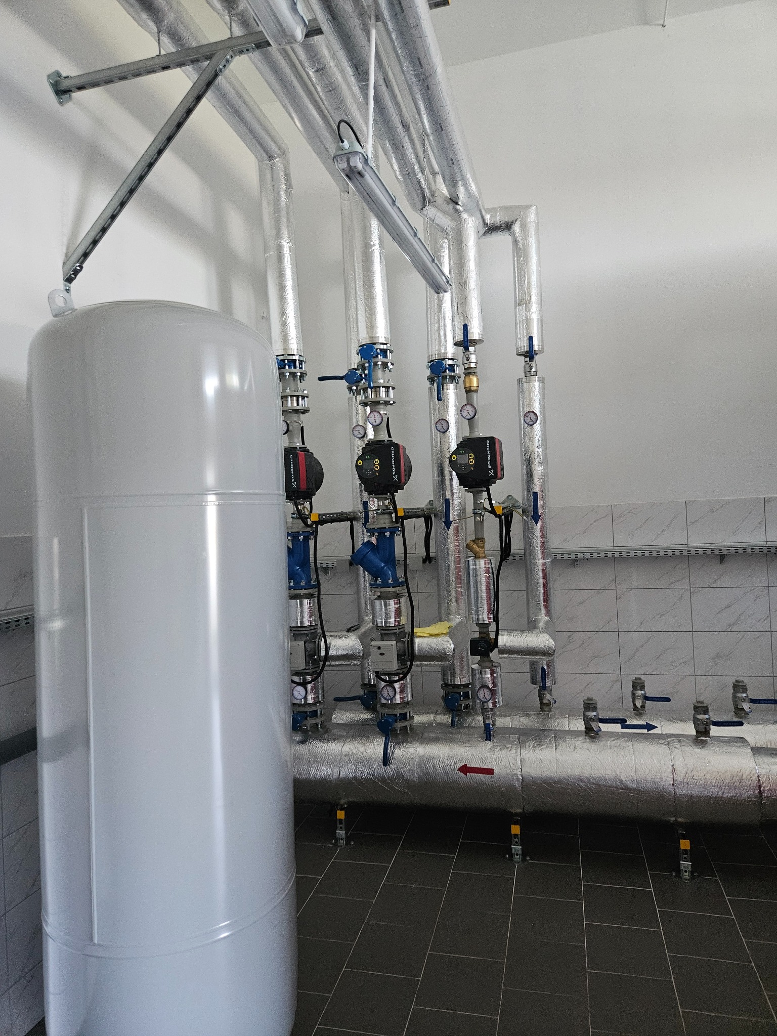 Instalacja hydrauliczna w pomieszczeniu technicznym: biały zbiornik buforowy, izolowane rury z zaworami i manometrami, pompy obiegowe Grundfos, szara podłoga z płytek, ściany wyłożone kafelkami.