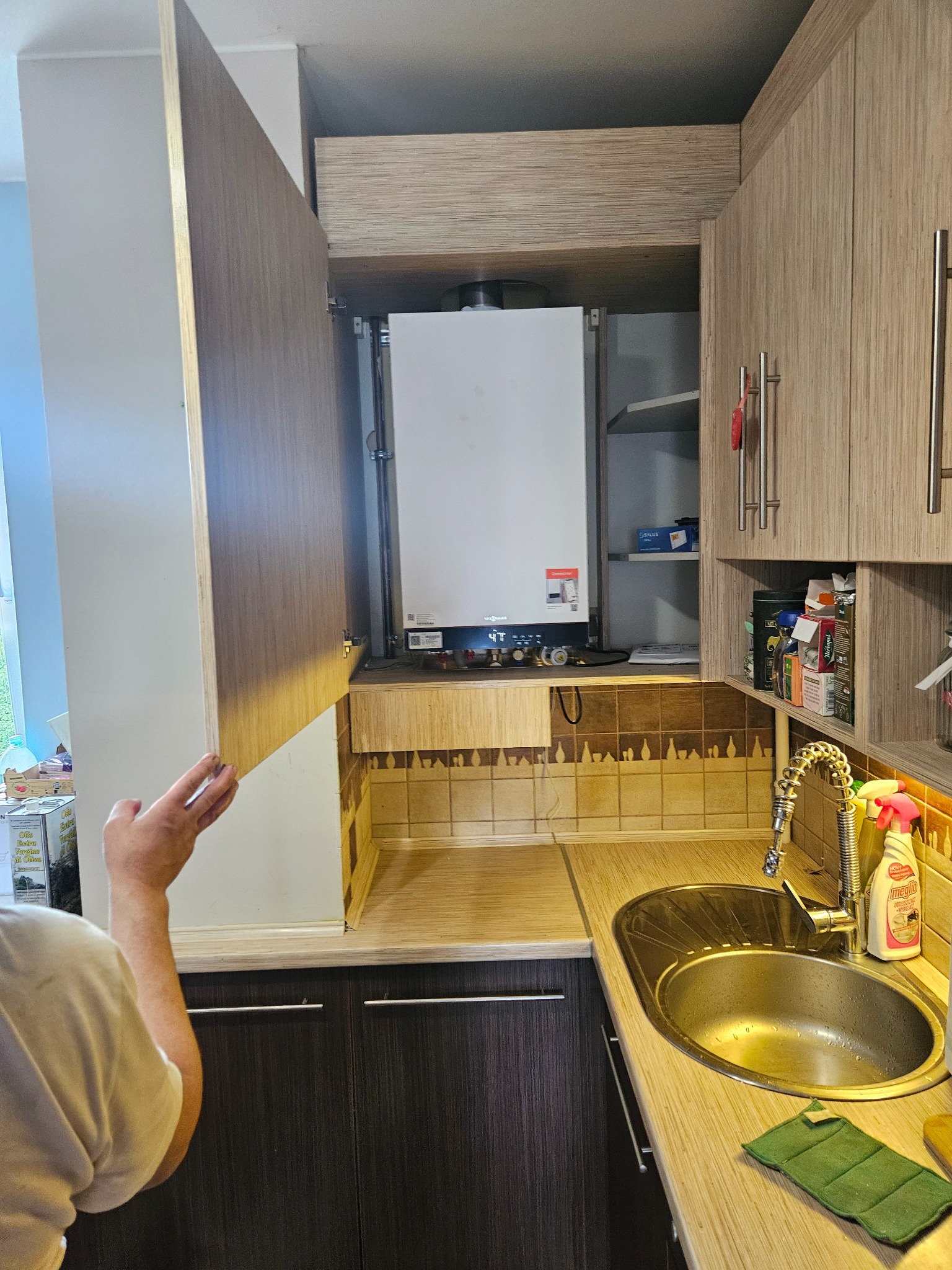 Piec gazowy schowany w szafce kuchennej z otwartymi drzwiczkami w Bielawie. Widoczny fragment instalacji i armatury. Ujęcie z bliska, w tle okno.