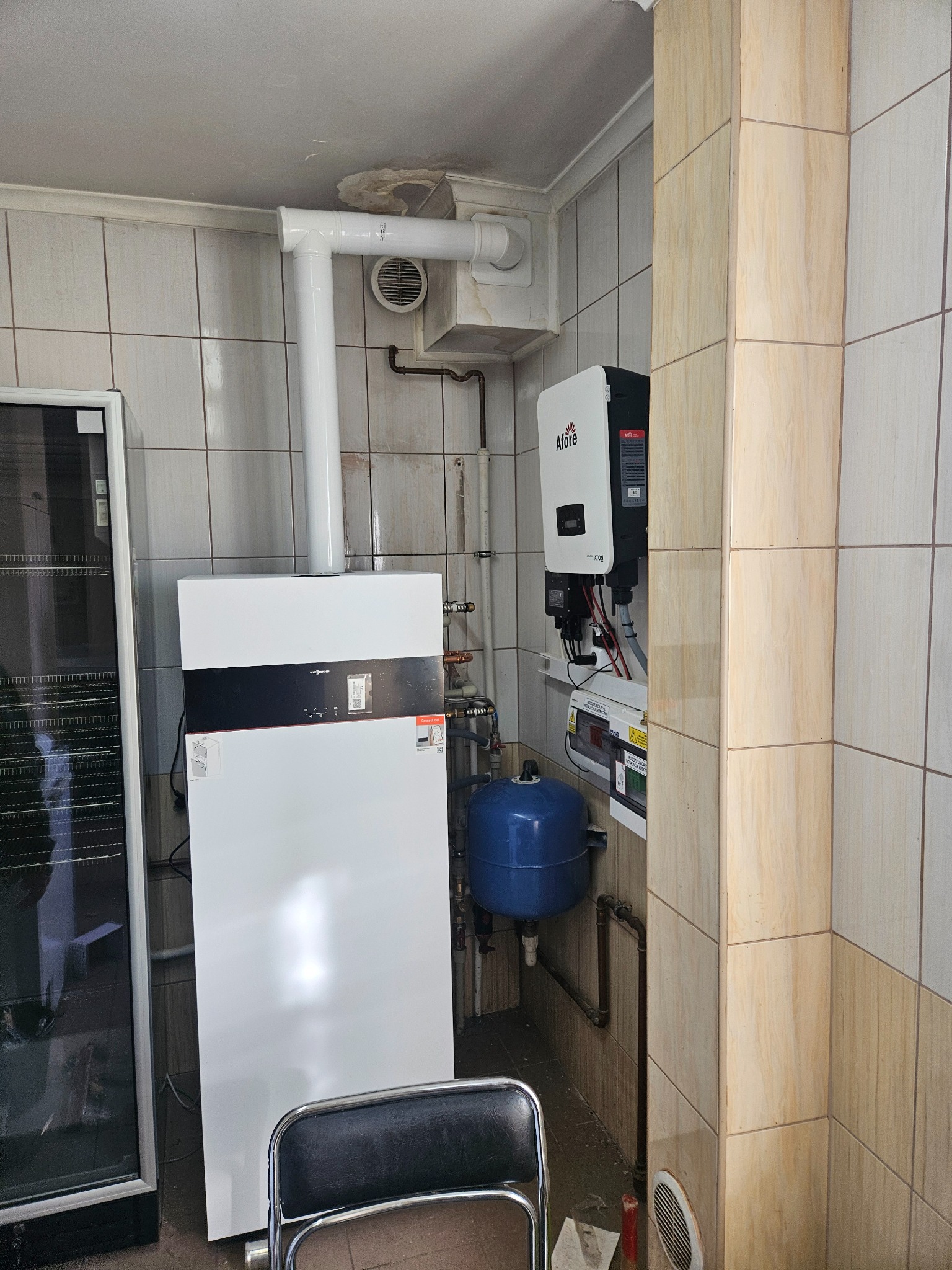 Biały piec Viessmann z rurą odprowadzającą, obok inwerter Afore na kafelkowanej ścianie. Niebieski zbiornik wyrównawczy i fragment krzesła w dolnej części kadru.