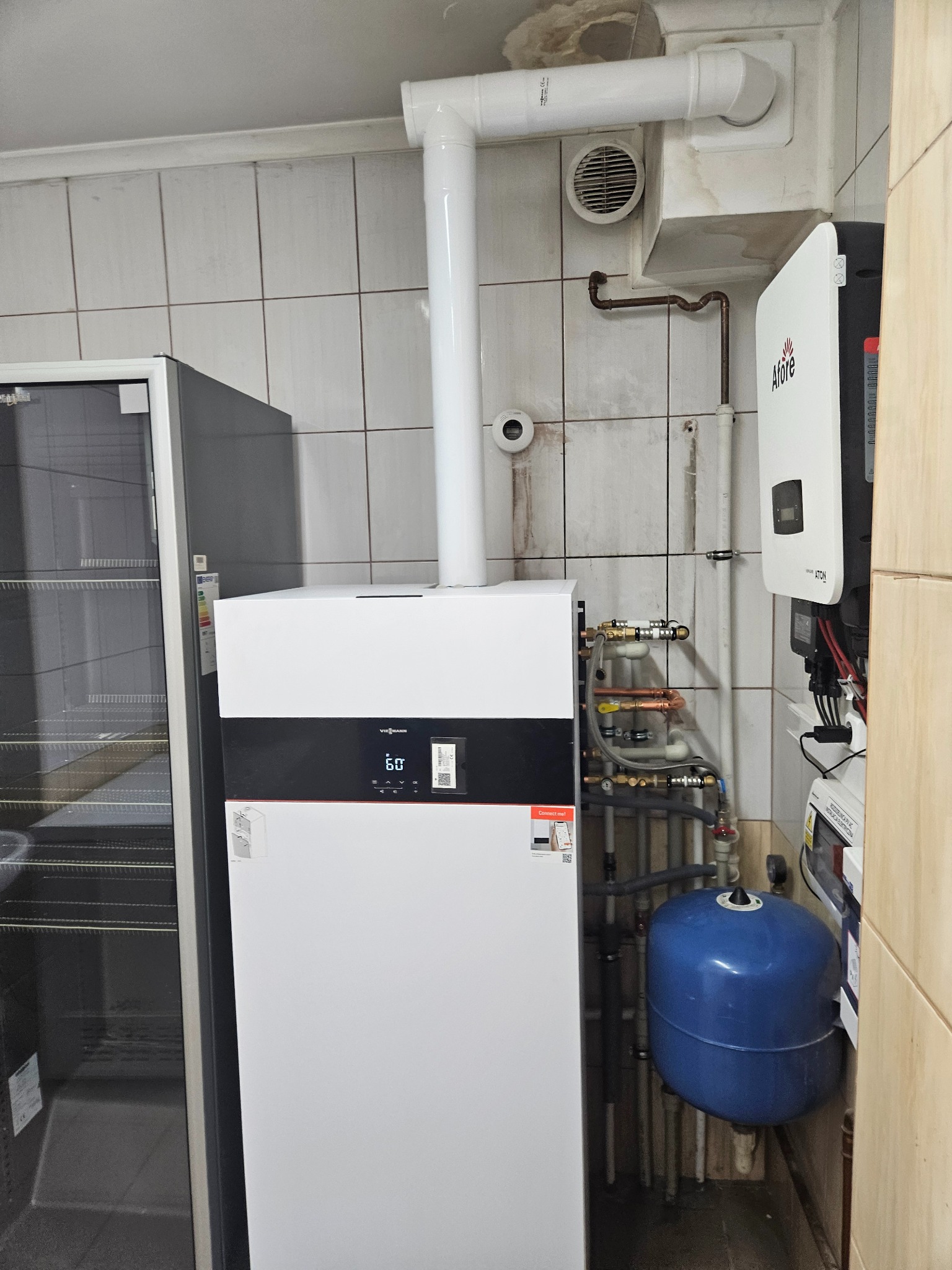 Biały piec Viessmann z czarnym panelem, rurami i niebieskim zbiornikiem wyrównawczym w kotłowni z białymi płytkami. Widoczny rekuperator Afore i wentylacja.