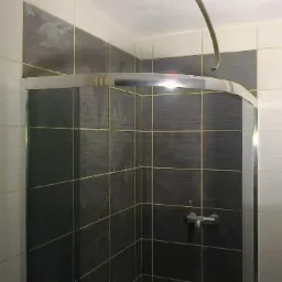 Wykończona kabina prysznicowa z szarymi i białymi płytkami w regularnym układzie. Widoczna chromowana bateria i fragment rurki. Całość w jasnym oświetleniu.