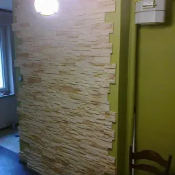 Ściana wykończona dekoracyjną okładziną imitującą kamień, oświetlona lampą sufitową. Widoczny fragment okna i zielona ściana z licznikiem gazu.