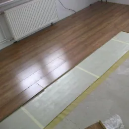 Układanie paneli podłogowych w jasnym wnętrzu. Częściowo ułożone panele w odcieniach brązu, widoczny podkład i surowa betonowa podłoga. Grzejnik na ścianie.