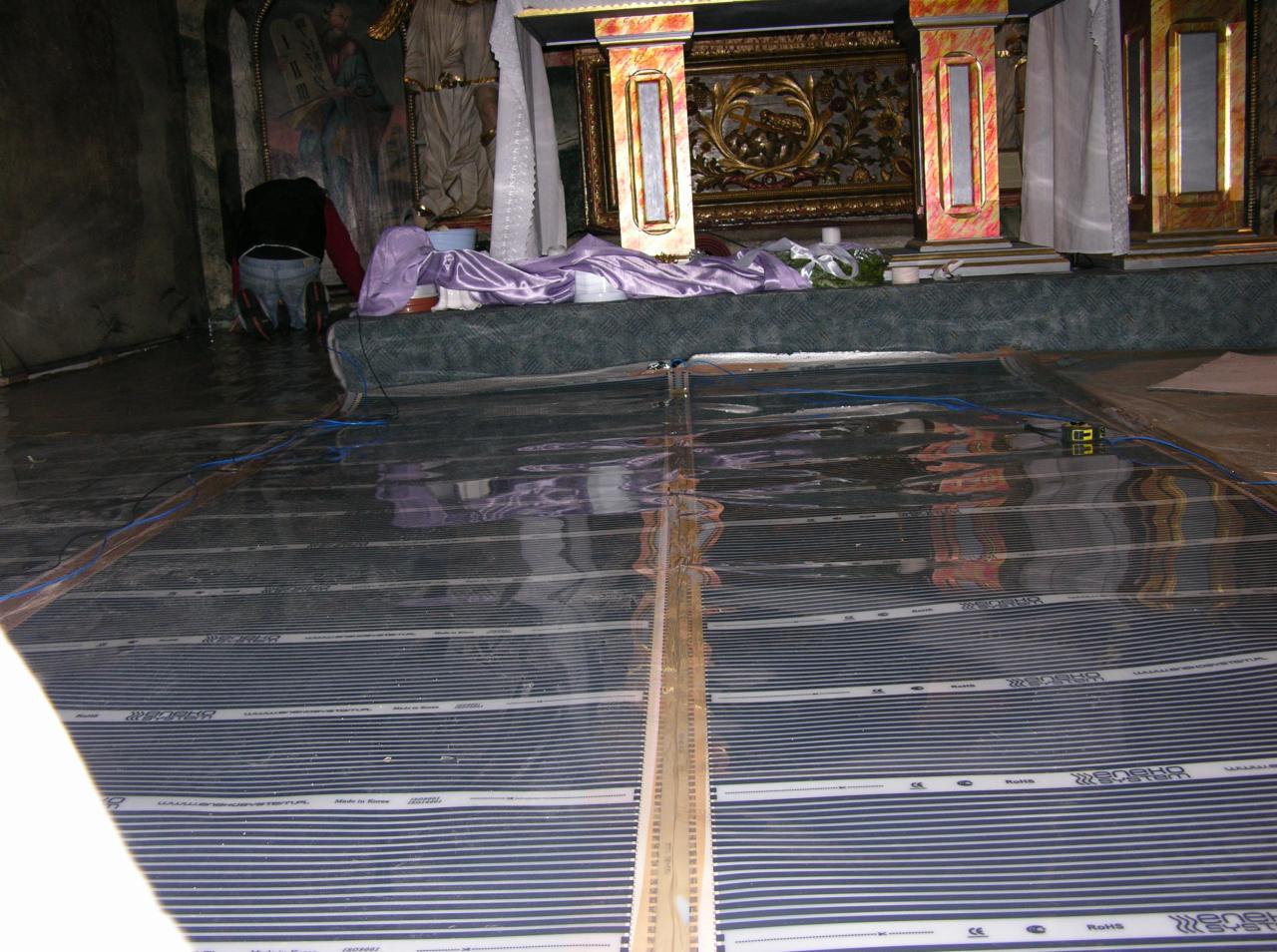 Montaż folii grzewczej Enenko System w zabytkowym wnętrzu. Widoczna osoba instalująca system, fragment ołtarza i elementy dekoracyjne.