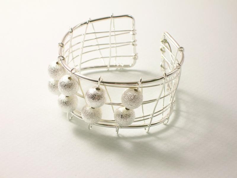 Srebrna bransoletka typu cuff z ażurową konstrukcją i wiszącymi, teksturowanymi kulkami, prezentowana na jasnym tle w neutralnym oświetleniu.