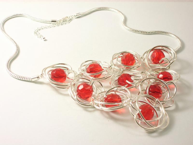 Srebrny naszyjnik z czerwonymi koralikami, artystycznie oplecionymi drutem, leżący na białym tle. Minimalistyczny design z wyraźnym akcentem kolorystycznym.