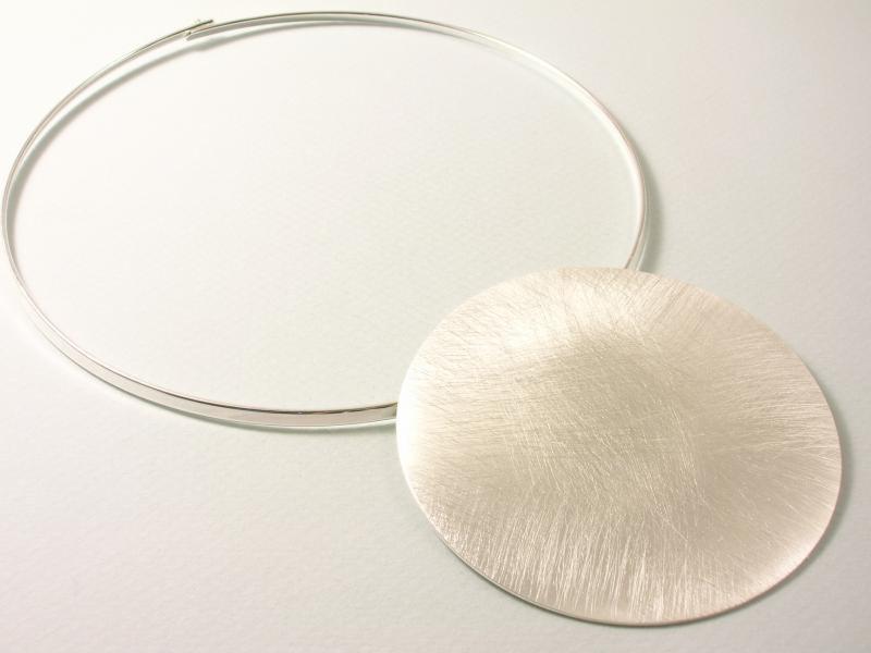Srebrny naszyjnik typu choker z dużą, okrągłą zawieszką, leżący na białym tle. Minimalistyczny design, widoczne delikatne rysy na powierzchni zawieszki.