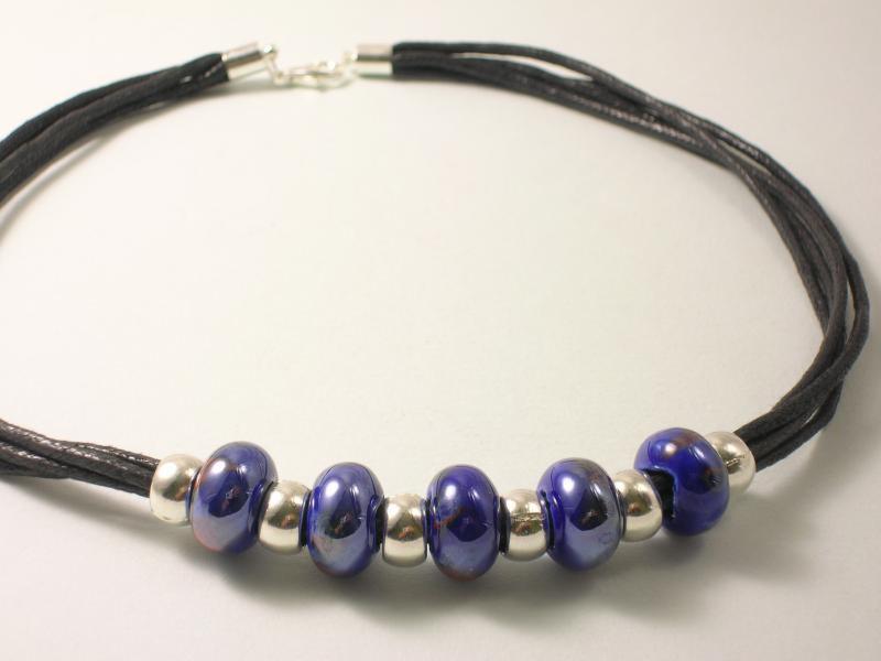 Elegancki naszyjnik z ciemnoniebieskimi, ceramicznymi koralikami i srebrnymi przekładkami na czarnym, skórzanym rzemyku, zapinany na srebrny karabińczyk.