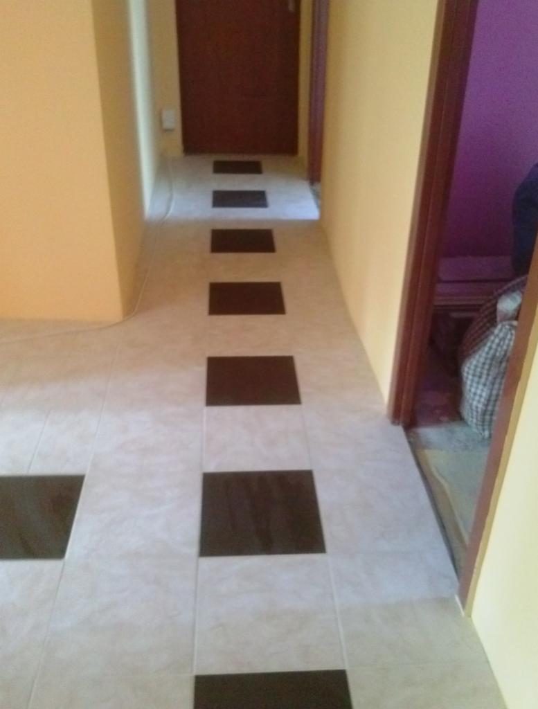 Beżowa podłoga z kwadratowymi, brązowymi wstawkami, tworzącymi wzór ścieżki, w korytarzu z żółtymi ścianami i brązowymi drzwiami.