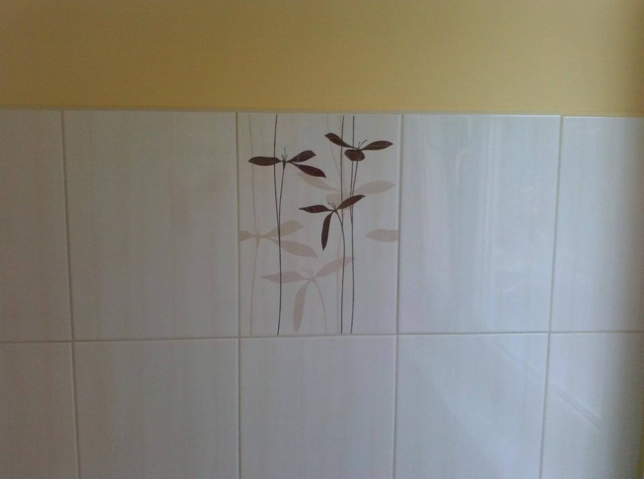 Białe, błyszczące płytki ścienne z motywem roślinnym, ułożone w rzędach, z żółtym pasem ściany u góry. Widoczne fugi i odbicia światła.