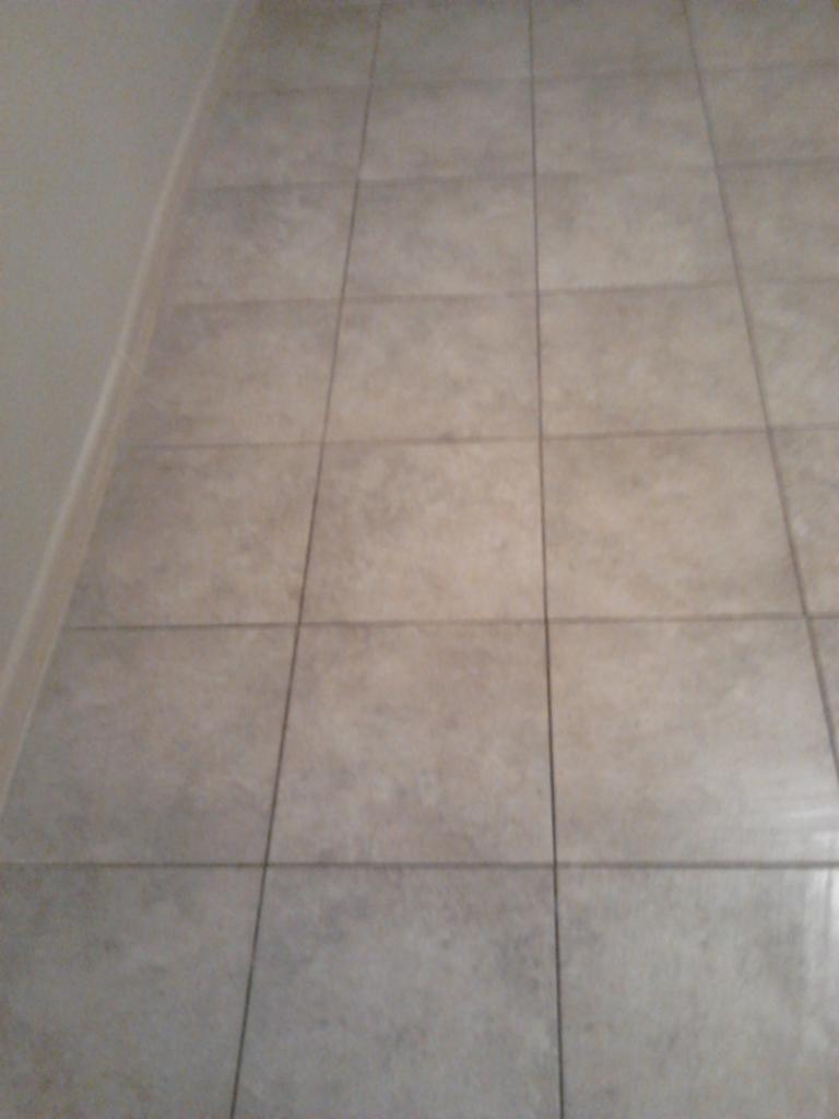 Beżowe, kwadratowe płytki podłogowe ułożone równo w rzędach, widoczne z perspektywy. Minimalistyczny design, szare fugi, fragment ściany po lewej stronie.