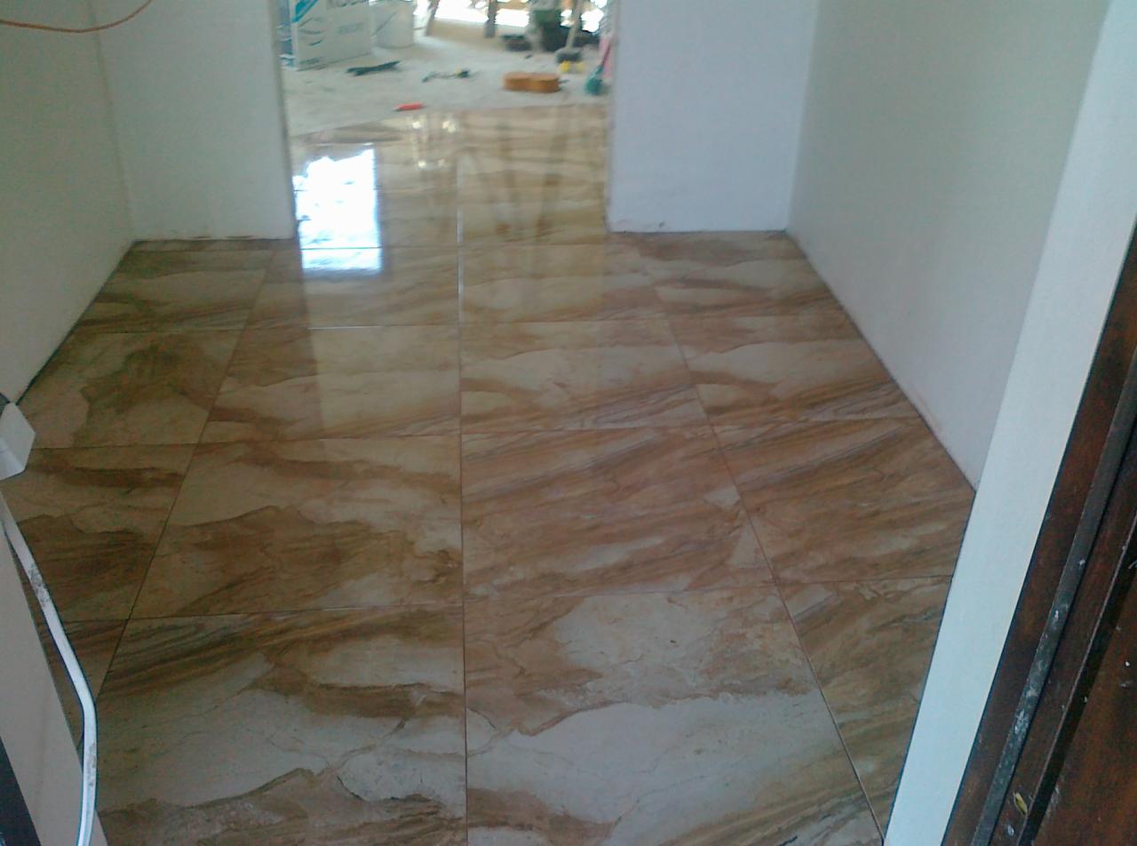 Ułożona, błyszcząca podłoga z płytek imitujących marmur w odcieniach beżu i brązu. Widoczny fragment ściany i drzwi. W tle nieporządek budowlany.