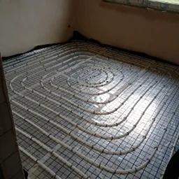 Białe rury ogrzewania podłogowego ułożone spiralnie na czarnej siatce w jasnym pomieszczeniu z oknem