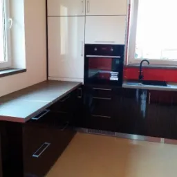 Nowoczesna kuchnia z białymi szafkami górnymi i ciemnobrązowymi dolnymi, wbudowany piekarnik, szary blat i czerwona mozaika nad zlewem.