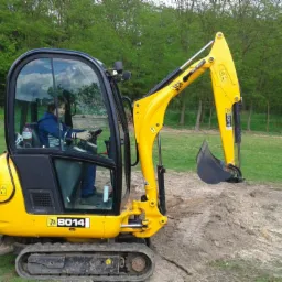 Żółta mini koparka JCB 8014 z operatorem w kabinie, pracuje na trawiastym terenie, z widocznym wykopem ziemi. Maszyna w trakcie niwelacji terenu.