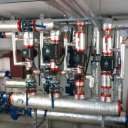 Złożona instalacja grzewcza z izolowanymi rurami, pompami obiegowymi Grundfos i manometrami, zamontowana w kotłowni.