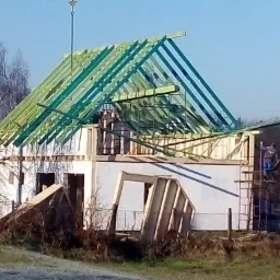 Budowa domu jednorodzinnego w stanie surowym otwartym z widoczną konstrukcją dachu z zielonych łat i kontrłat, pracownik na rusztowaniu oraz elementy szalunków na pierwszym planie.