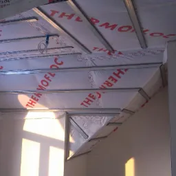 Poddasze z widoczną izolacją Thermofloc, metalowymi profilami i oknem łukowym, w trakcie prac wykończeniowych, z widocznym oświetleniem słonecznym wpadającym przez okno.