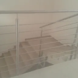 Schody wewnętrzne z metalową, minimalistyczną balustradą z cienkich, poziomych prętów, widok z góry na jasne stopnie i białe ściany.