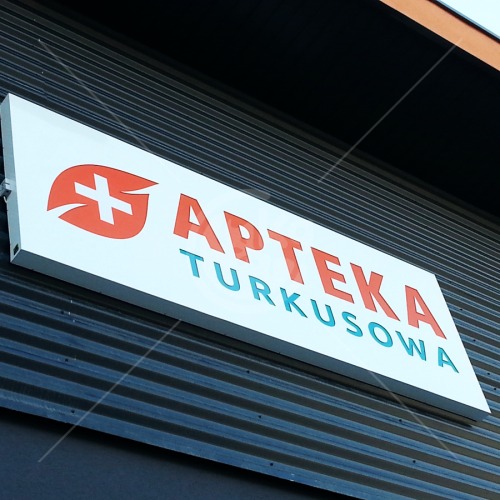 Szyld apteki 'Apteka Turkusowa' z logo w postaci czerwonego krzyża w liściu, zamontowany na ciemnej, metalowej elewacji budynku.
