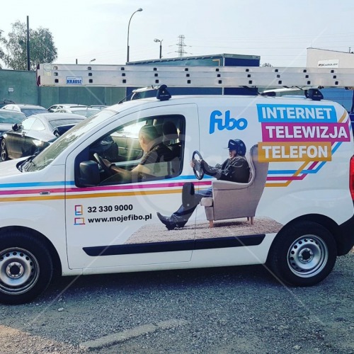 Biała furgonetka z oklejeniem reklamowym firmy telekomunikacyjnej w Czeladzi, przedstawiającym kierowcę w fotelu i hasła 'Internet, Telewizja, Telefon'. Na dachu drabina.