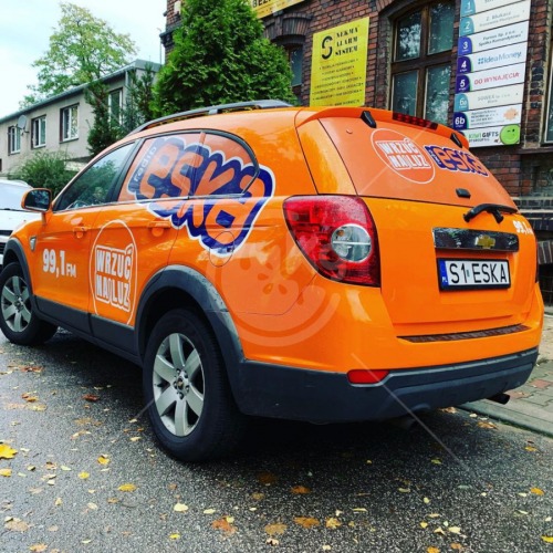Pomarańczowy samochód Chevrolet Captiva z oklejeniem reklamowym Radia Eska, zaparkowany na ulicy z liśćmi.