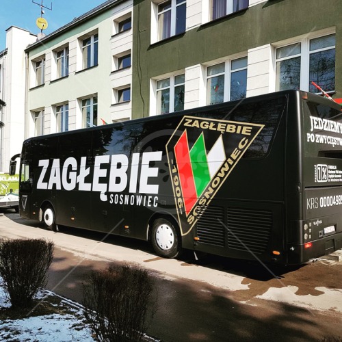 Czarny autobus z dużym białym napisem 'ZAGŁĘBIE SOSNOWIEC' i herbem klubu sportowego w kształcie rombu z zielono-biało-czerwonymi pasami, zaparkowany na ulicy przy budynku.