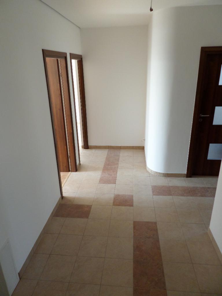 Jasne wnętrze z beżową podłogą w kwadraty z brązowymi akcentami. Widoczne drzwi z drewnianymi ościeżnicami i zaokrąglona ściana. Minimalistyczny design.