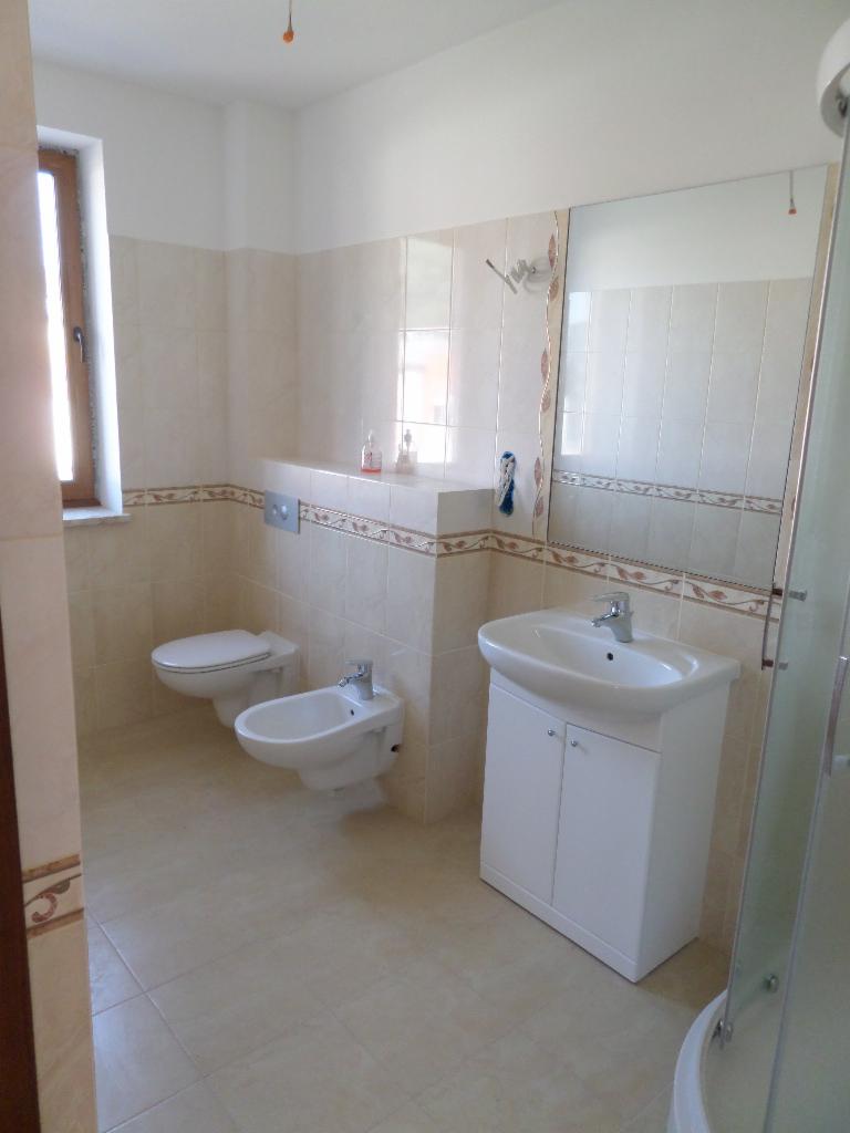 Jasna łazienka po remoncie: beżowe płytki z dekoracyjnym pasem, biała umywalka z szafką, toaleta i bidet. Widoczne lustro i fragment kabiny prysznicowej.