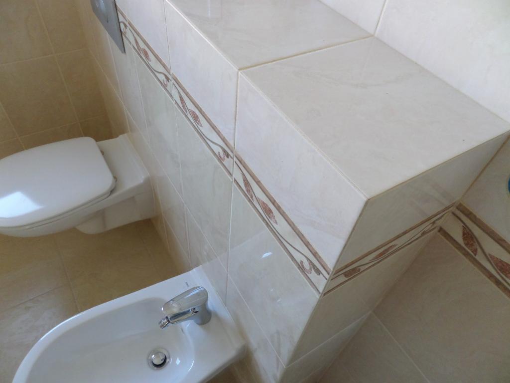 Jasna łazienka z toaletą i bidetem. Płytki ceramiczne z motywem roślinnym zdobią ściany i obudowę stelaża podtynkowego. Ujęcie z góry, widoczne elementy armatury.