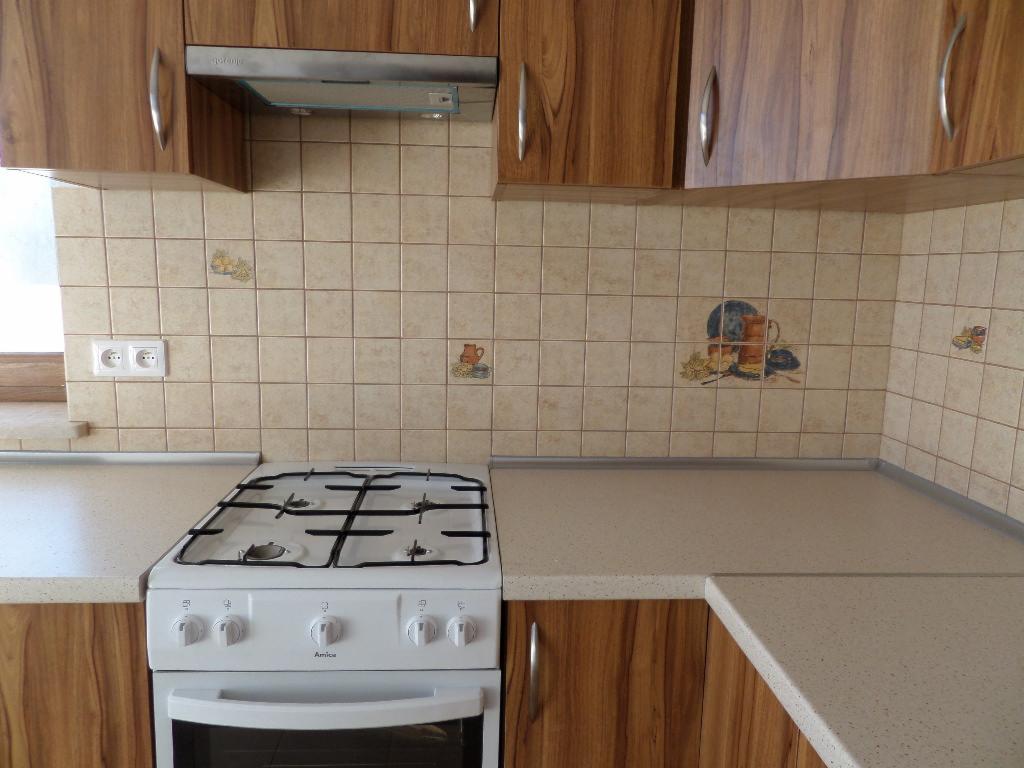 Kuchnia z drewnianymi szafkami, beżowymi płytkami z motywami i białą kuchenką Amica. Ujęcie z perspektywy użytkownika, widoczny fragment okna z lewej.